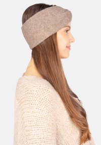 Un bandeau en tissu beige doux, avec un design torsadé. La texture semble confortable, idéale pour se tenir au chaud par temps froid.