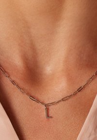 Zilveren ketting met ovale schakels en een kleine, bevestigd "L" hanger. Soepele textuur op de huid. Eenvoudig, modern ontwerp.