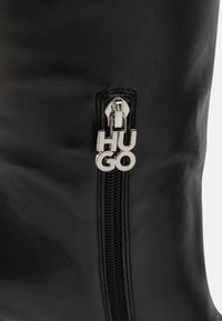 Czarny skórzany but zasilany srebrnym zamkiem błyskawicznym ozdobionym logo "HUGO" oraz teksturowanym materiałem. Zamek biegnie pionowo wzdłuż boku.