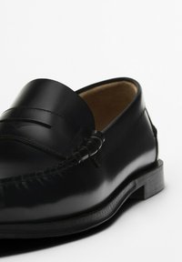 Mocassino in pelle nera con finitura liscia, presenta una punta arrotondata, dettagli cuciti e un tacco basso impilato. All'interno ha una fodera in pelle color cuoio.