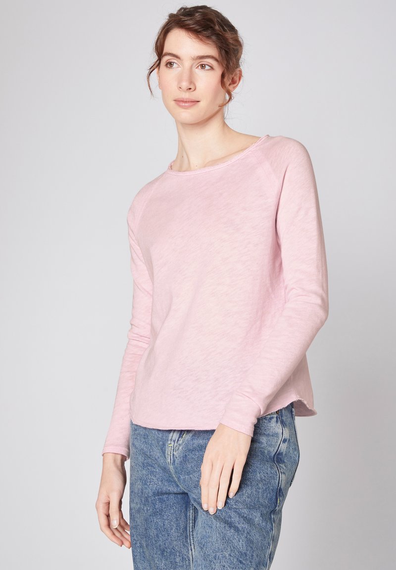 Helles rosa Langarmhemd aus einem weichen, strukturierten Stoff. Entspannte Passform mit Rundhalsausschnitt und leicht abgerundetem Saum. Getragen mit blauen Jeans.