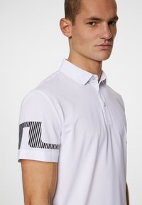 Weißes Poloshirt mit Kragen, aus strukturiertem Stoff und einem schwarzen gestreiften Logo auf dem linken Ärmel.