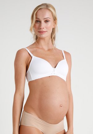 Gravid kvinne som har på seg en hvit bh og beige undertøy, står mot en lys, ensfarget bakgrunn og smiler milde.