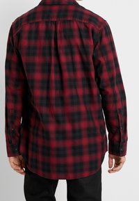 Man som bär en långärmad skjorta i röd och svart rutigt mönster med knäppta manschetter, sedd bakifrån mot en enfärgad bakgrund.