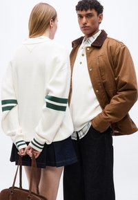 Άνδρας με καφέ μπουφάν και λευκό πουλόβερ κοιτάει μπροστά· γυναίκα με λευκό πουλόβερ με πράσινες ρίγες κρατάει καφέ τσάντα πίσω από την πλάτη της.