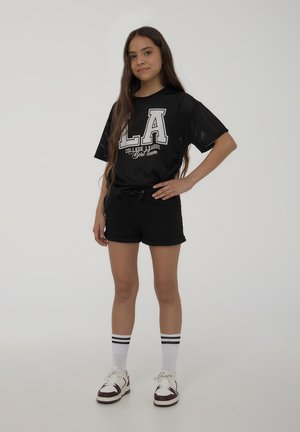 T-shirt nera in rete con grafica "LA", abbinata a pantaloni neri. Calzini bianchi con strisce nere e sneakers bianche con accenti bordeaux.