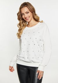 Witte sweatshirt met zilveren en witte versieringen, ronde halslijn en lange raglanmouwen. Gecombineerd met zwarte leren broeken.