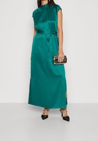 Teal satin maxiklänning med korta ärmar, hög hals och sprund på sidorna. Kombinerad med svarta spetsiga klackskor och en liten metallisk handväska.