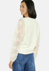 Maglione crema con maniche a sbuffo in pizzo caratterizzate da motivi floreali; corpo aderente con orlo a coste. Abbinato a jeans in denim blu.