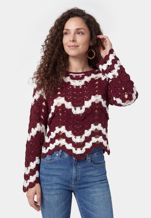 Vrouw met krullend haar draagt grote ringoorbellen, een bordeauxrode en witte gehaakte trui en blauwe spijkerbroek, raakt haar haar aan en kijkt weg.