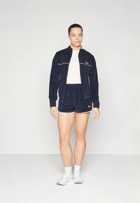 Marineblauw velours trainingspak met witte accenten, bestaande uit een jack met ritssluiting en bijpassende korte broek. Ontworpen met een casual pasvorm en eenvoudige lijnen.