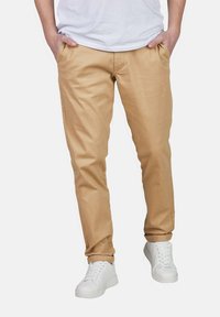 Khaki Chinos mit schmaler Passform, ausgestattet mit Seitentaschen und Gürtelschlaufen, kombiniert mit weißen Sneakers. Glatte Textur und neutrale Farbe.