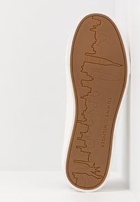 Białe sneakersy z teksturowaną brązową gumową podeszwą, na której znajdują się wyryte wizerunki panoramy miasta, w tym Statua Wolności oraz różne budynki.