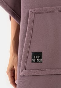 Fioletowy polarowy ponczo z dużą przednią kieszenią, widocznymi szwami oraz czarną metką z napisem „MON AMI” w dolnym rogu.