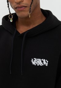 Sudadera negra con capucha que presenta un bolsillo tipo canguro en la parte delantera, capucha con cordones y un logotipo "VISION" bordado en blanco en el pecho. Tejido suave y texturizado.