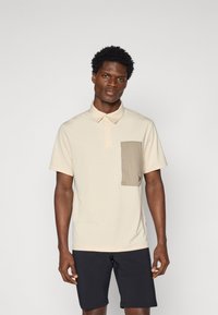 Camisa polo bege com um bolso no peito texturizado em bege, mangas curtas e gola. Usada com calções escuros. Tecido liso.