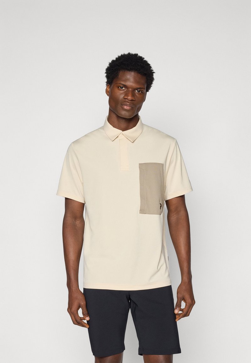 Camisa polo bege com um bolso no peito texturizado em bege, mangas curtas e gola. Usada com calções escuros. Tecido liso.