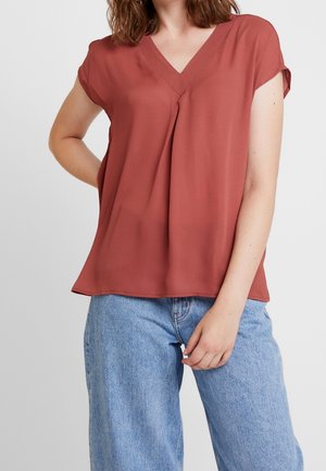 Femme portant un chemisier à manches courtes col en V couleur rouille et un jean en denim bleu clair, debout, une main sur la hanche, l'autre détendue.