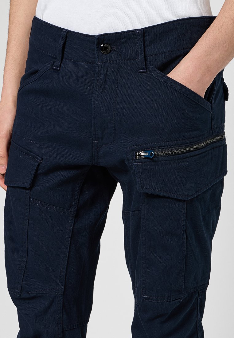 Navy Cargo-Hosen aus Baumwollmischung, ausgestattet mit mehreren Taschen, einer Reißverschlusstasche mit blauen Akzenten und einem Knopfverschluss an der Taille.