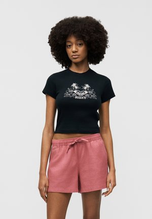 Göndör hajú nő fekete, pálmafás mintájú crop topban és rózsaszín megkötős rövidnadrágban, sima háttér előtt áll.