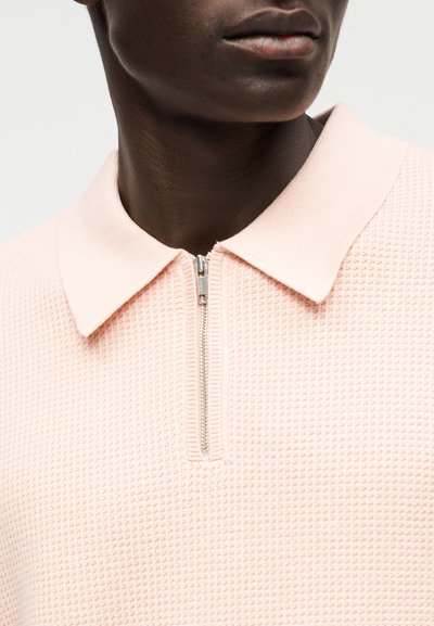 Κοντινό πλάνο ενός ατόμου που φοράει ένα ροζ ανοιχτό υφασμάτινο πουκάμισο με φερμουάρ και γιακά, εστιάζοντας στον γιακά και την περιοχή του φερμουάρ.