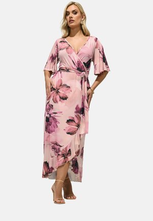 Kvinde iført en lyserød blomsterprint slå-om-kjole med flæsekant og flagrende ærmer, accessoriseret med guldbøjler, armbånd og beige hæle.
