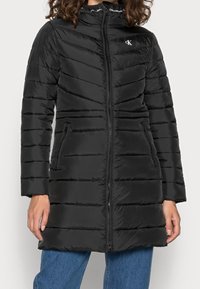 Manteau en duvet noir avec fermeture éclair à l'avant, design en chevrons matelassé, deux poches latérales et col droit. Tissu lisse et léger.