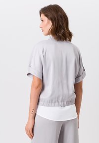 Helles graues Blusenshirt mit lockerer Passform und hochgerollten Ärmeln, das eine geraffte Taille aufweist. Darunter ist eine weiße Schicht sichtbar. Glatte Textur.