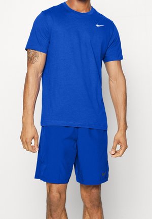 Mand iført blå Nike t-shirt og matchende blå shorts, der står mod en ensfarvet baggrund med synlige arm-tatoveringer.
