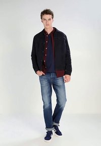 Giovane uomo in piedi con le mani in tasca, indossa una giacca scura, una camicia bordeaux, una t-shirt blu navy, jeans arrotolati e sneakers blu navy, contro uno sfondo neutro.