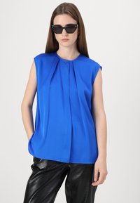 Top senza maniche blu con un fronte plissettato e scollo rotondo, realizzato in tessuto liscia. Indossato con pantaloni in pelle nera con un motivo testurizzato.
