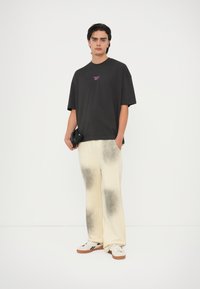 Camiseta negra oversized con logo morado, pantalones crema con patrón de salpicaduras negras y zapatillas blancas con acentos negros.