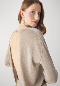 Pull beige métallisé à dos ouvert, avec une texture douce et une coupe ample, présentant une fermeture dans le dos et un tissu scintillant.
