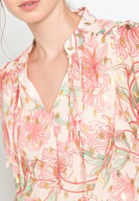 Lekka, przezroczysta bluzka w jasnoróżowym kolorze z kwiecistymi wzorami w kolorze koralowym, zielonym i złotych akcentach, z dekoltem w kształcie litery V i falbaniastym kołnierzykiem.