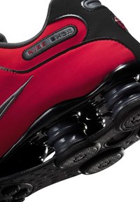 Nike Shox-Sneaker verfügen über ein rotes und schwarzes Obermaterial aus synthetischem Material, eine schwarze Gummisohle und markante stoßdämpfende Säulen unter der Ferse.