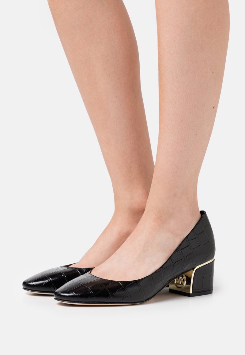 Chaussures noires à talon bloc bas à motif crocodile portées pieds nus, présentant un détail doré métallique dans le design du talon.