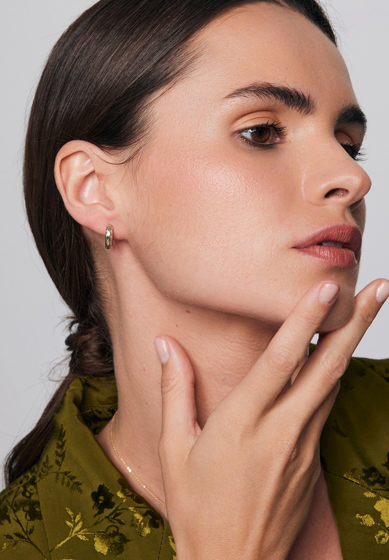 Złota kolczyk w hoop z pokręconą teksturą, noszony na lewym uchu. Modelka ma ciemne włosy, nosi zieloną kwiecistą bluzkę, ma jasną skórę i naturalne paznokcie.