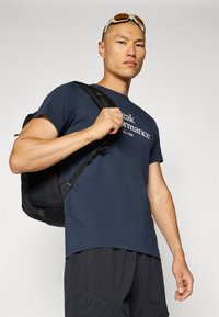 Camiseta de color navy con el logo "Peak Performance", combinada con una mochila negra. El modelo muestra un agarre fuerte en la correa de la mochila, llevando gafas de sol.