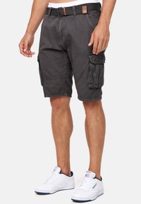 Homme portant un short cargo gris foncé avec poches latérales, une ceinture noire et des baskets blanches Reebok sur un fond uni.