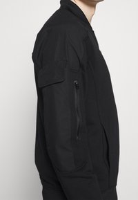 Černá bomber bunda s kapsou na zip na levém rukávu, žebrovaným límcem a manžetami a hladkou texturou.