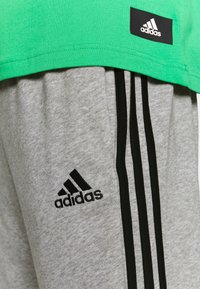 Camiseta de algodón verde combinada con pantalones de chándal grises con rayas negras y un logo de Adidas bordado en negro en el muslo.