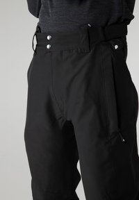 Čierne vonkajšie nohavice vyrobené z odolného materiálu, s uzatvárateľnou vreckom na zips, dvoma kovovými cvokmi na páse a elegantným designom.