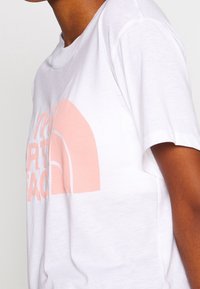 Bílá bavlněná tričko s velkým broskvovým logem a zakřivenými tvary. Krátké rukávy a kulatý výstřih dotvářejí design.