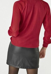 Rotes Langarm-Oberteil mit gerippter Textur an den Ärmeln, kombiniert mit einem schwarzen Leder-Minirock. Das Oberteil ist weich und hat eine glatte Oberfläche.