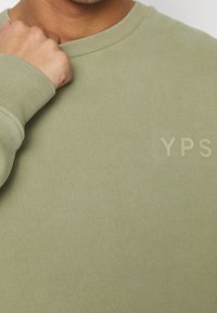 Olivově zelená mikina s kulatým výstřihem, žebrovanými manžetami a měkkou texturou. Má decentní logo "YPS" ve stejné barvě.