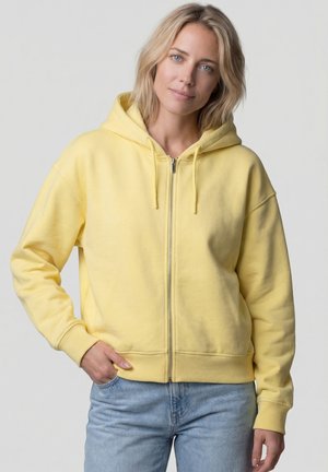 Blonde Frau mit kurzen Haaren, die eine gelbe Kapuzenjacke mit Reißverschluss und hellblaue Jeans trägt, steht mit einer Hand in der Tasche vor einem schlichten Hintergrund.