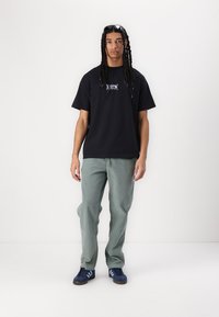 Tricou din bumbac negru cu detalii grafice, pantaloni texturiți de culoare verde deschis și adidași albăstrii cu dungi albe. Modelul adoptă o poziție neutră.