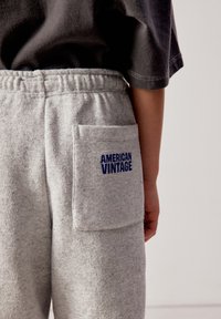 Enfant portant un pantalon de survêtement gris clair avec taille élastique et grande poche marquée "AMERICAN VINTAGE" en texte bleu, t-shirt noir à manches courtes.