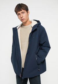 Giacca zip-up blu navy con un morbido rivestimento in pile beige e cappuccio, dotata di due tasche laterali e un piccolo dettaglio del logo sul petto.