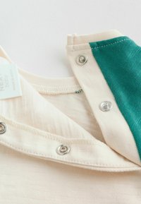 Nahaufnahme eines cremefarbenen und grünen Baby-Kleidungsstücks mit silbernen Druckknöpfen und einem Stoffetikett mit der Aufschrift "Next baby Made in India".